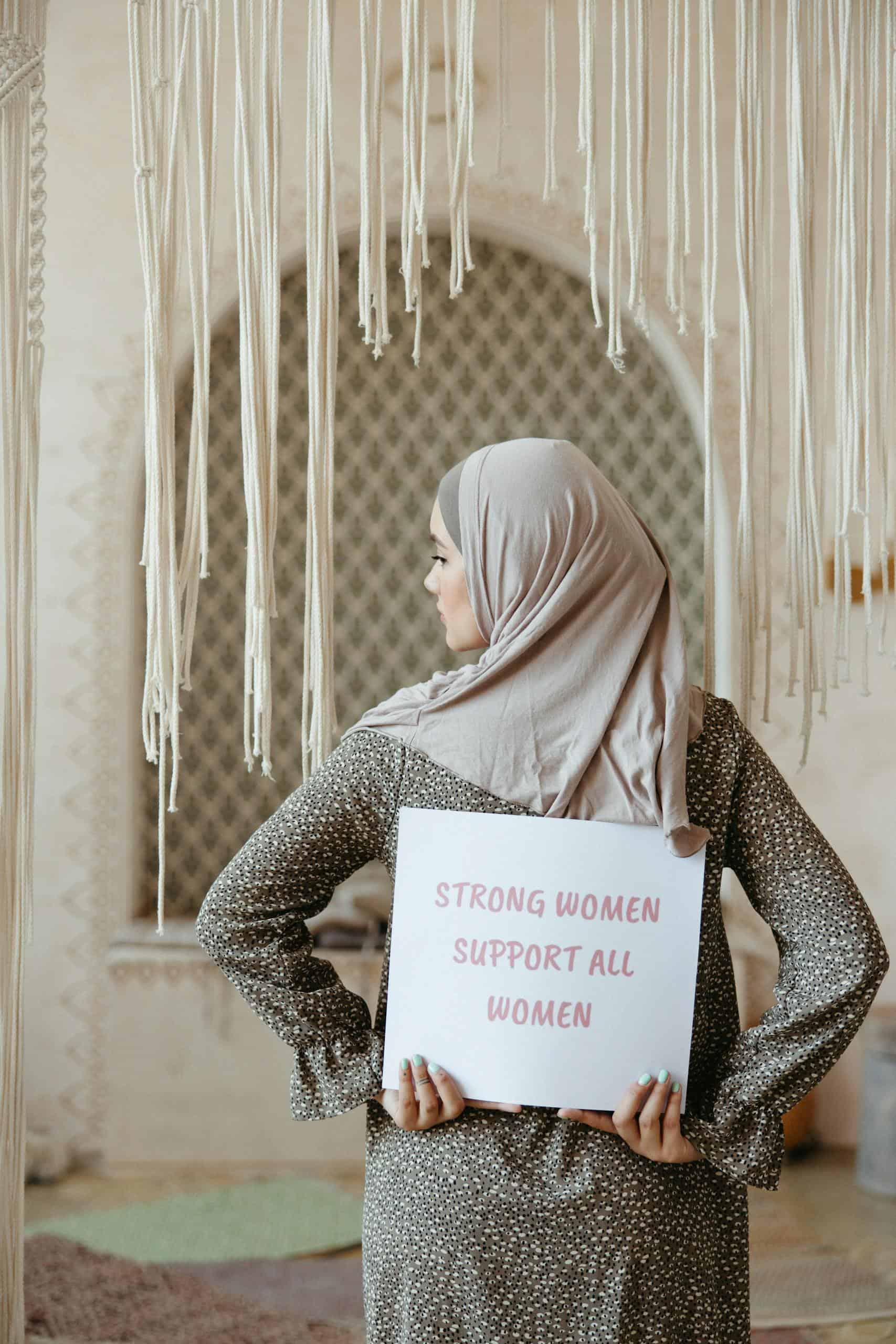 A Muslim woman in a hijab holds a sign with an empowering message, promoting unity and support.